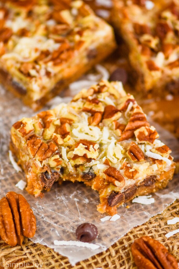 up close overhead picture of a seven layer bar on a piece of parchment paper with a bite missing, coconut shreds and pecan pieces visible on the top of the gooey bar