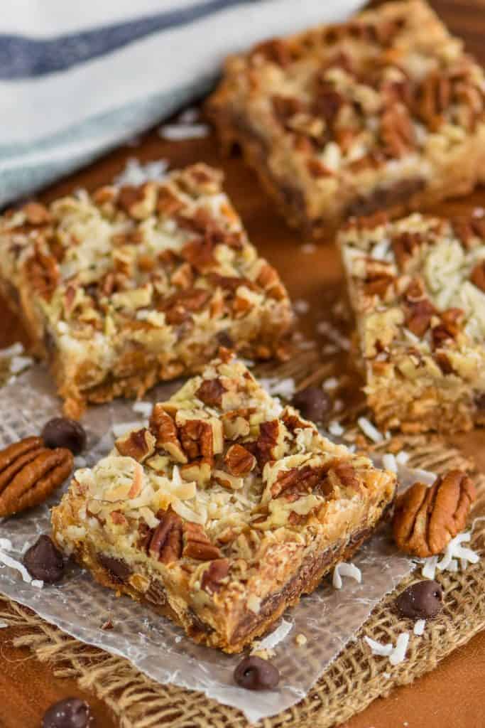 four seven layer bars sitting on parchment paper on a wood board with coconut, pecans, and chocolate chips around them