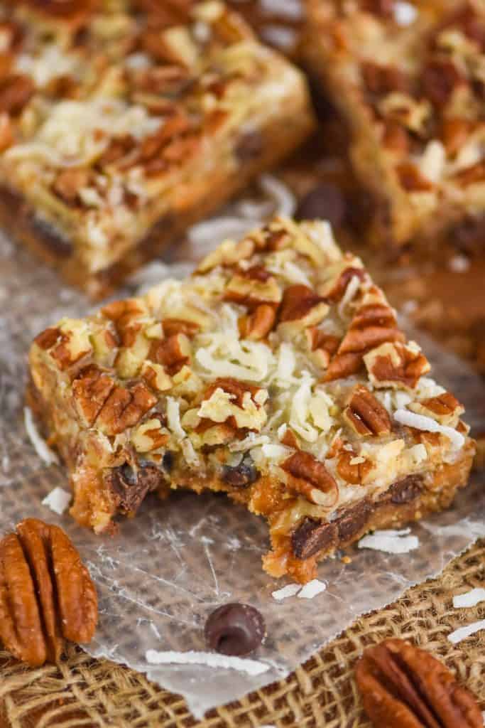 up close overhead picture of a seven layer bar on a piece of parchment paper with a bite missing, coconut shreds and pecan pieces visible on the top of the gooey bar