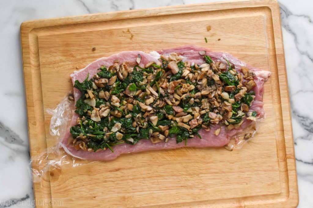 a flattened pork loin on a cutting board that is covered in spinach and mushrooms filling