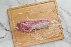 a pork tenderloin that has been trimmed of excess fat and is on a sheet of Saran Wrap on a cutting board