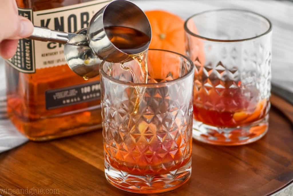 stainless steal double jigger pouring bourbon whiskey into a small tumbler to make an old fashioned cocktail