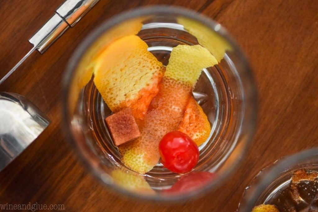 an overhead close up view of the bottom of a tumbler glass with a lemon peel, orange peel, maraschino cherry, and sugar cube soaked with brown bitters