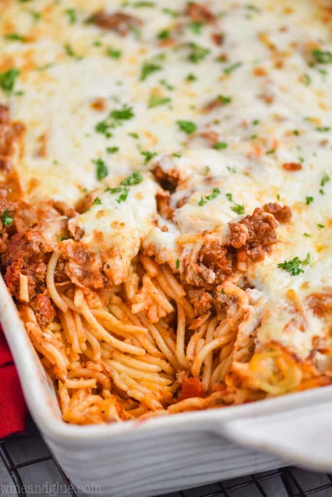 a close up of a white ceramic baking dish full of baked spaghetti topped with cheese and fresh parsley