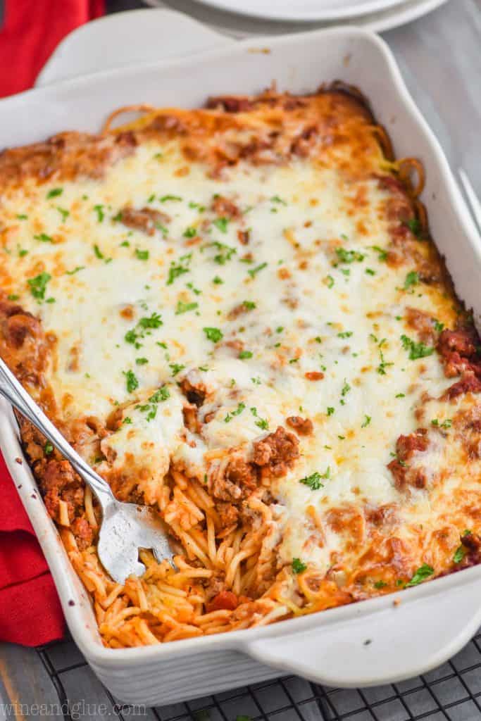 pulled back view of baked spaghetti casserole in a baking dish, topped with cheese and fresh parsley