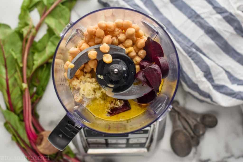 overhead view of a food processor with roasted beets, chick peas, garlic, tahini, lemon juice, and olive oil