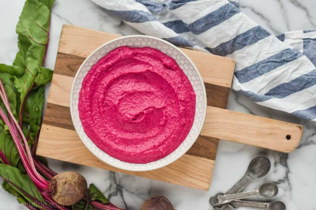 a white bowl full of beet lemon hummus on a wood cutting board with a striped cloth napkin next it and whole raw beets with their stems and leaves on the other side