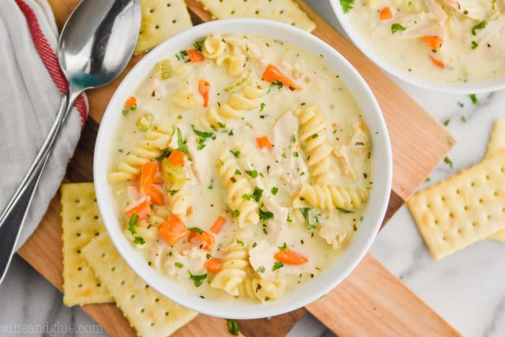 overhead view of a bowl of creamy chicken noodle soup recipe, garnished with fresh parsley, lots of fresh parsley visible, on a wooden cutting board, crackers around the bowl