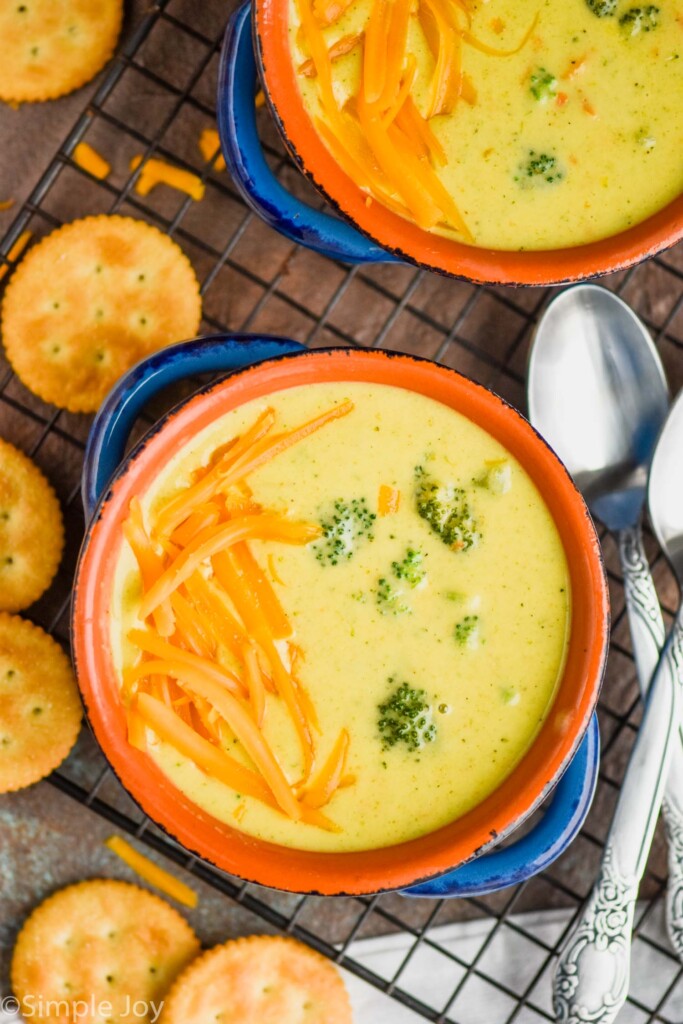 overhead view of a broccoli cheddar soup recipe in two small blue soup bowls on a wire cooling rack, with round orange crackers next to it and two spoons on the side