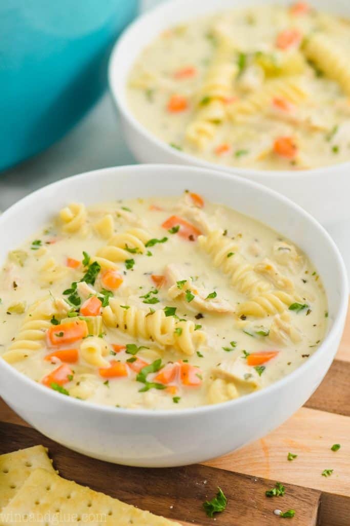 a small white bowl of creamy chicken noodle soup on a cutting board with another bowl behind, both garnished with fresh parsley