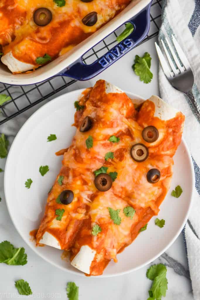 two easy beef enchiladas sitting on a white plate, garnished with black olive slices and fresh cilantro next to a cooling rack that has a baking dish on it and a fork next to it