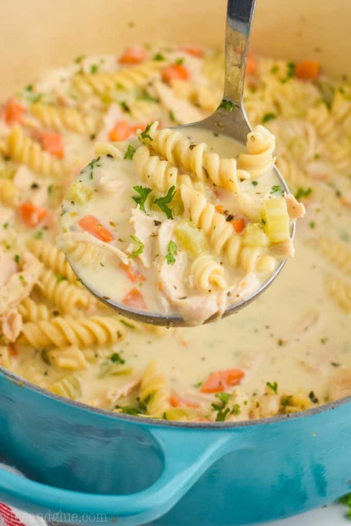 a ladle pulling a creamy chicken noodle soup out of a teal stock pot, made with spiral rotini noodles