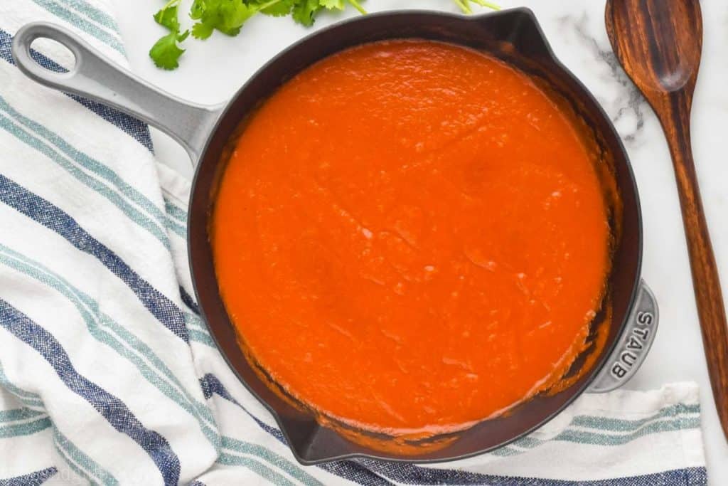 overhead of a gray skillet on a marble counter with enchilada sauce recipe, next to a white and blue striped cloth napkin and fresh cilantro