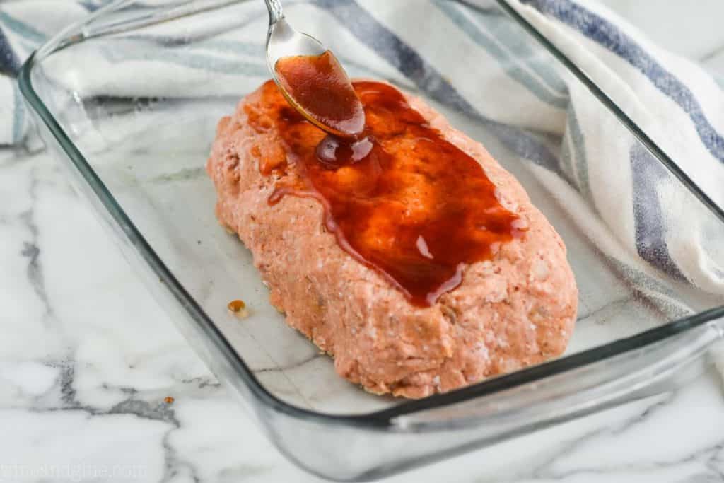a turkey meatloaf in a glass baking dish that hasn't been cooked yet and is having bbq sauce spooned on top