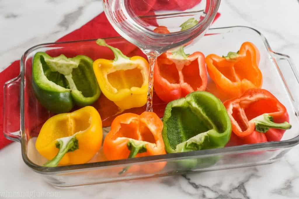 a glass baking dish full of 8 half bell peppers that are empty and face up, with water being carefully poured into the dish