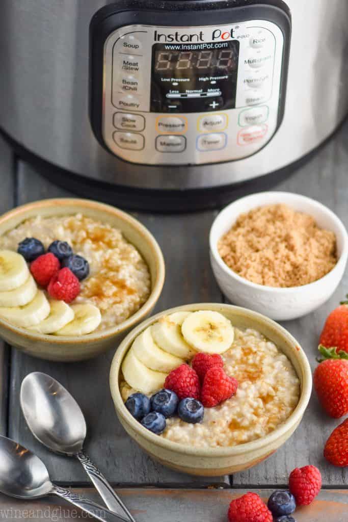 two small bowls of instant pot steel cut oats recipe garnished with berries and banana slices next to a small bowl of brown sugar with an instant pot in the background