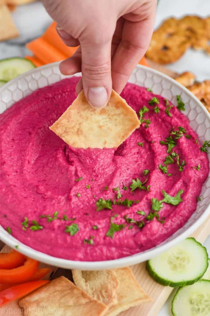 a hand dipping a pita chip into a bowl of roasted beet hummus garnished with fresh parsley