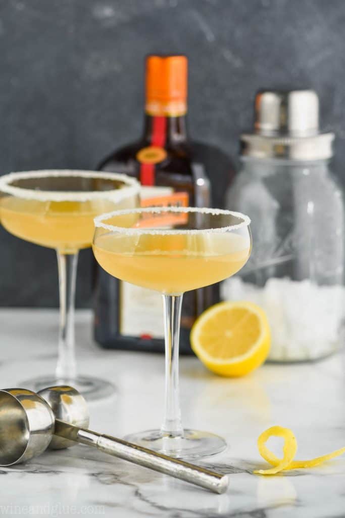 two coupe glasses rimmed with sugar holding a sidecar recipe with a double sided metal jigger in the foreground, a lemon twist off to the side and a bottle of cointreau and a cocktail shaker in the background