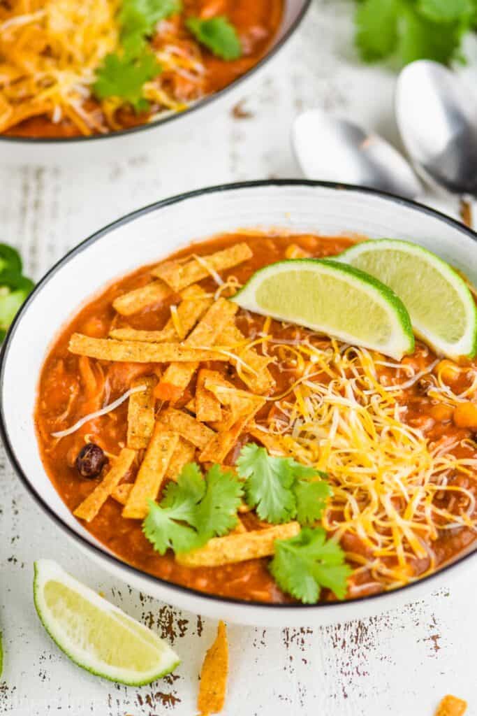 side view of a small white bowl with a dark rim containing chicken enchilada soup, topped with tortilla strips, lime wedges, cheese, and cilantro