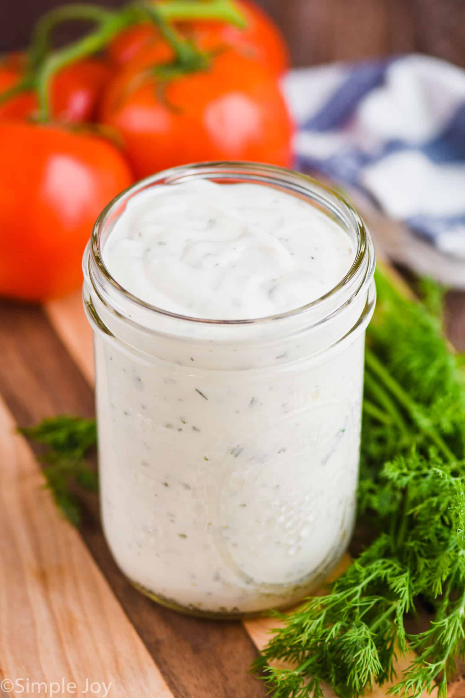 small mason jar of homemade ranch dressing on a cutting board next to fresh dill with tomatoes in the background