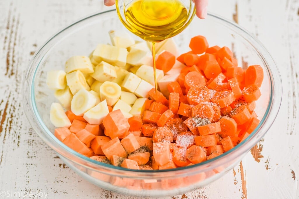 pouring olive oil into a large bowl full of raw cut up parsnips, sweet potatoes, and carrots