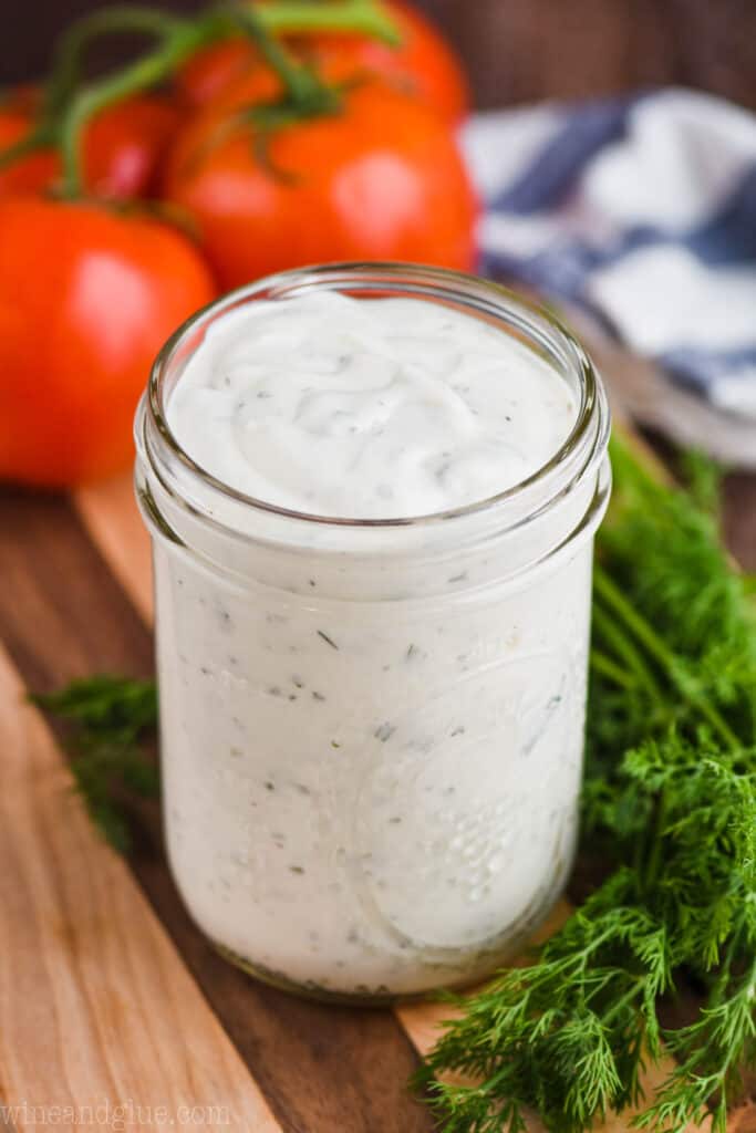 small mason jar of homemade ranch dressing on a cutting board next to fresh dill with tomatoes in the background