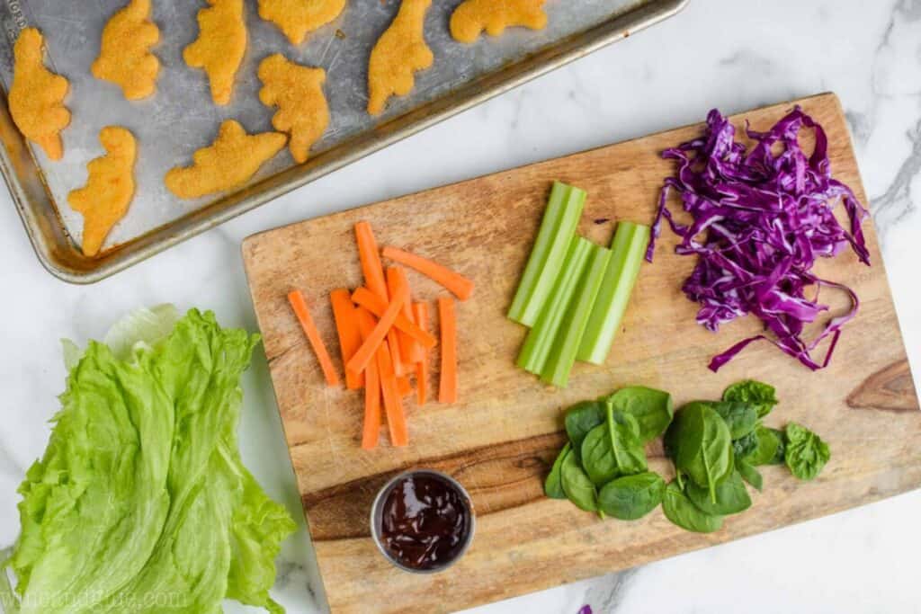 overhead view of ingredients for lettuce wraps for kids on a cutting board with Dinosaur chicken nuggets on a baking tray next to it