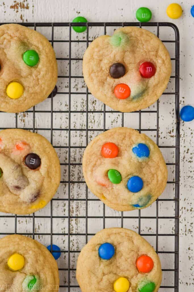 overhead photo of six m and m cookies on a wire cooling wrack on a white wood board with m & ms around them