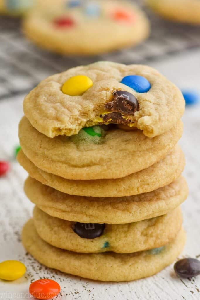 A stack of 6 m&m cookies. The first one in the stack has a small bite out of it. 