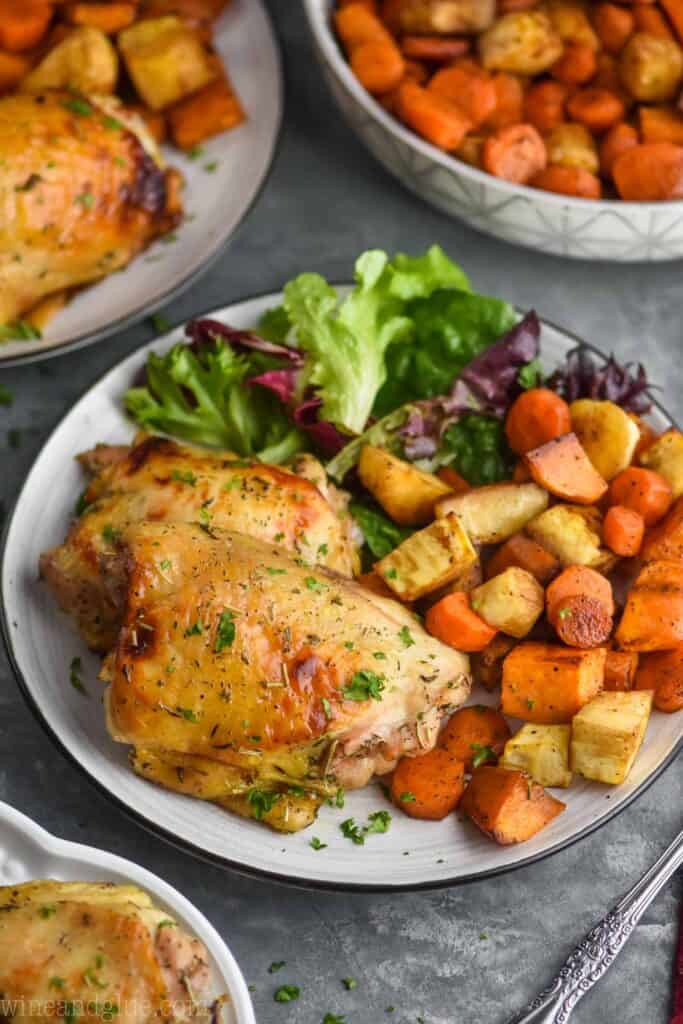 pulled back view of chicken thighs that have been marinated and baked on a plate with lettuce and roasted rooted vegetables