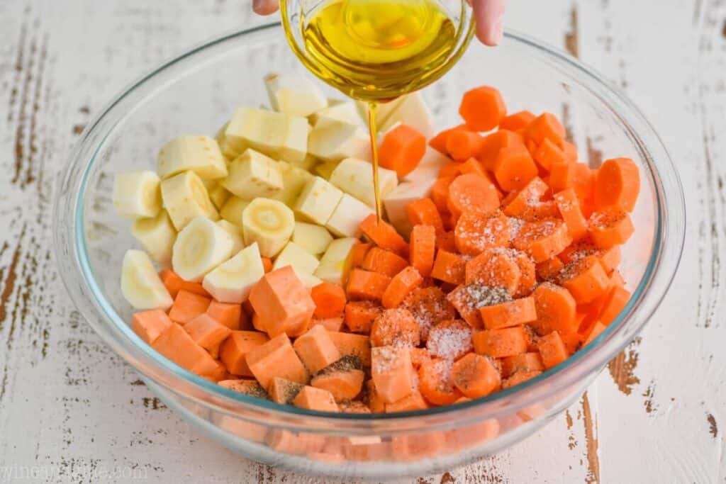 pouring olive oil into a large bowl full of raw cut up parsnips, sweet potatoes, and carrots