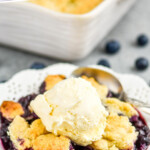pinterest graphic of close up of a white dessert plate with blueberry cobbler recipe topped with vanilla ice cream, served with a spoon, says: the best blueberry cobbler