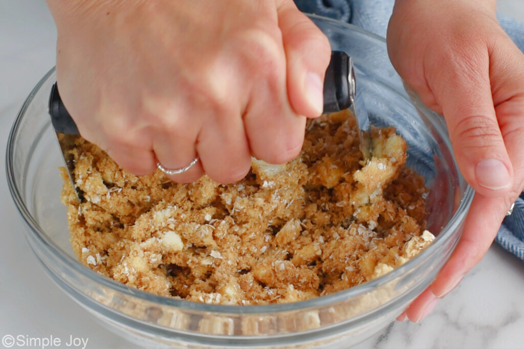 hands bringing together butter, brown sugar, and oats to with a pastry cutter to make French Toast Casserole