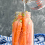 Clear mason jar filled with whole carrots. Measuring cup above jar pouring clear liquid into jar to make pickled carrots.