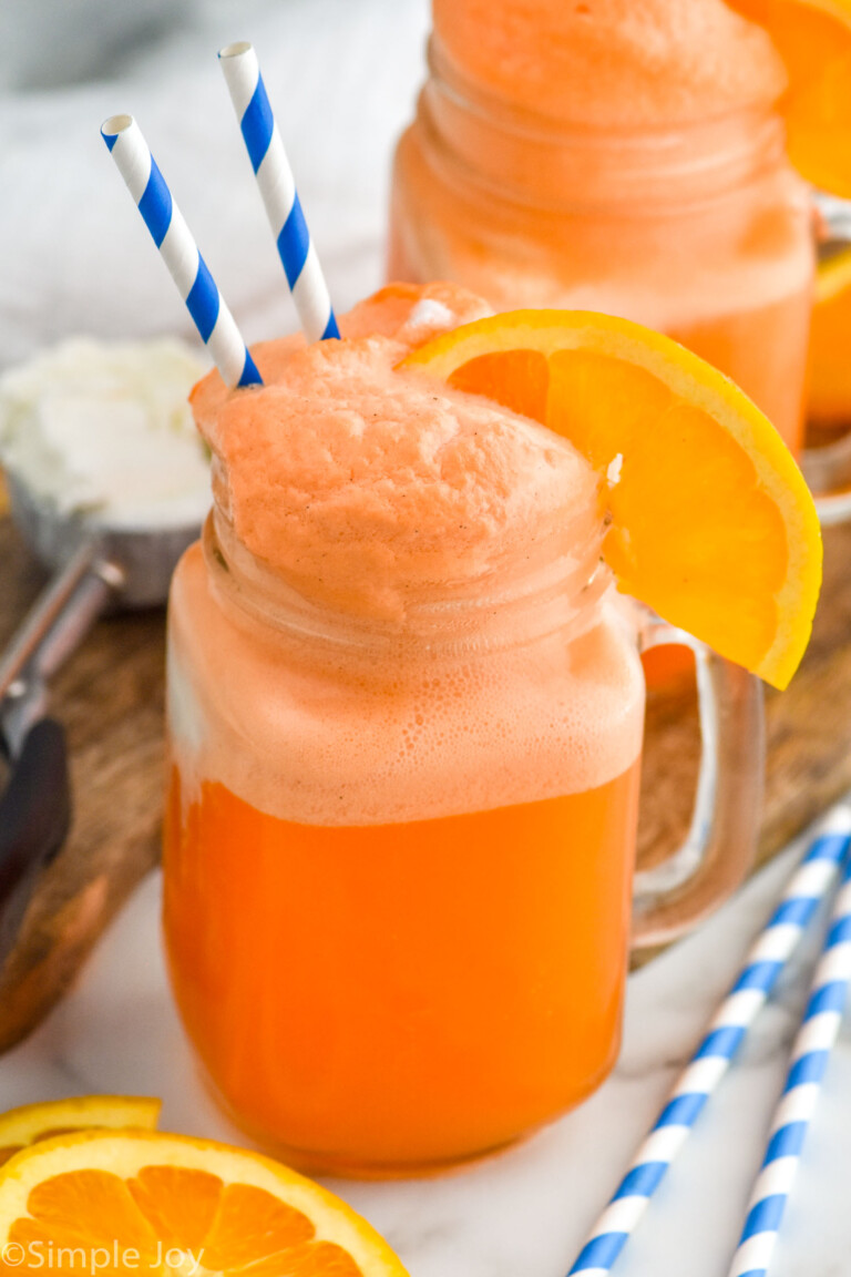 Side view of Orange Creamsicle served in a glass mug garnished with straws and an orange slice. Another glass of Orange Creamsicle on the counter, along with extra orange slices and straws.