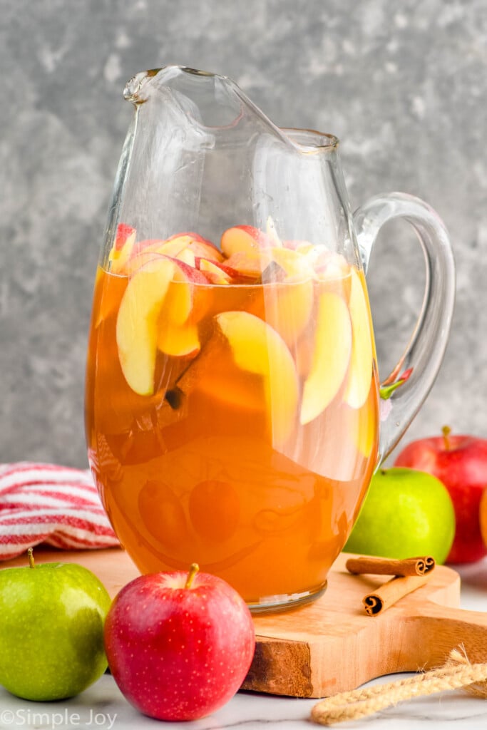 Side view of pitcher of Apple Cider Sangria with apples and cinnamon sticks beside it.