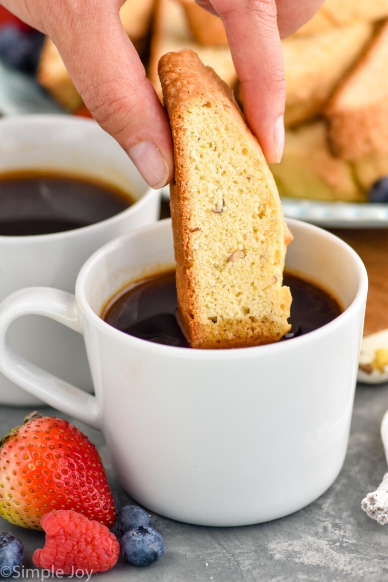 Photo of a person's hand dipping Biscotti in a cup of coffee. Cup of coffee, fruit, and plate of Biscotti beside.