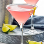 Photo of Cosmopolitan Cocktail with another cosmo and cocktail shaker behind. Lime wedges beside.