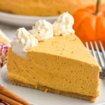 Photo of a slice of No Bake Pumpkin Cheesecake on a plate with pumpkin and cinnamon sticks beside. Whole No Bake Pumpkin Cheesecake behind.