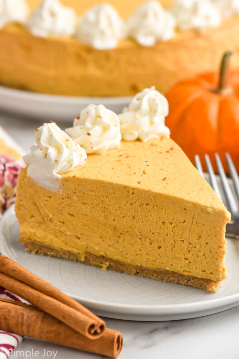 Photo of a slice of No Bake Pumpkin Cheesecake on a plate with pumpkin and cinnamon sticks beside. Whole No Bake Pumpkin Cheesecake behind.