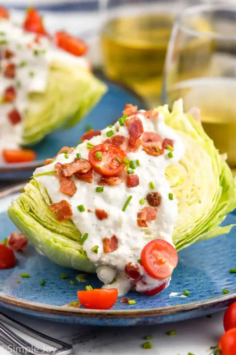 Wedge Salad served on a plate with tomatoes, bacon, and dressing. Another Wedge Salad and glasses of wine beside.