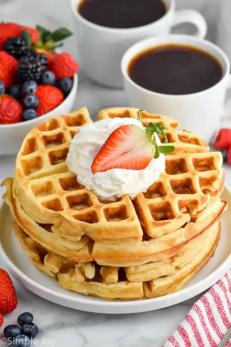 Stack of Buttermilk Waffles on a plate garnished with whipped cream and strawberry. Cups of coffee and bowl of berries beside.