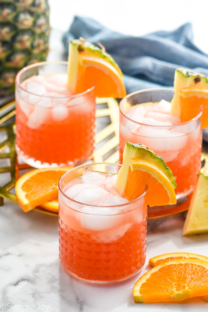 three glasses of rum punch with ice, garnished with orange slices and pineapple wedges