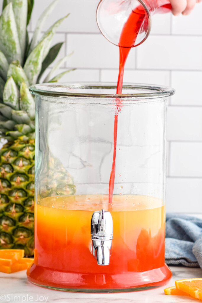 glass of grenadine pouring into a drink dispenser of rum punch ingredients to make rum punch. Pineapple and orange slices sitting in background