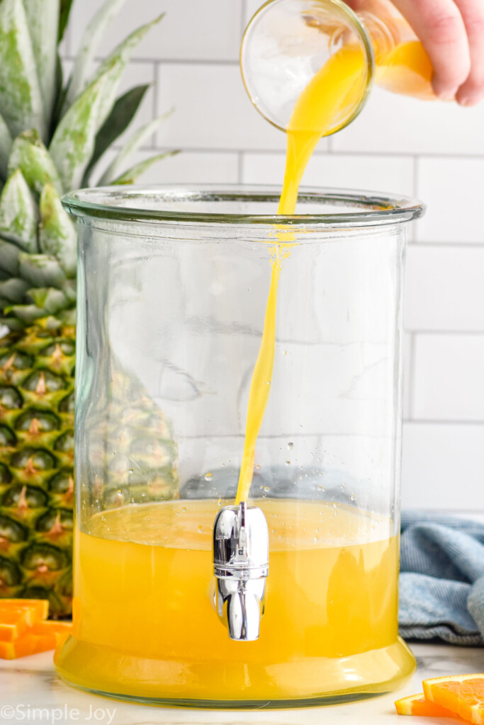 orange juice pouring into a drink dispenser to make rum punch. Fresh pineapple and orange slices sitting in background