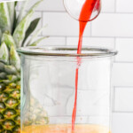 pinterest graphic for rum punch. Text says "the best rum punch recipe simplejoy.com" Image shows grenadine pouring into drink dispenser of rum punch ingredients. Fresh pineapple and orange slices sitting in background.