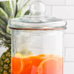 Pinterest image for rum punch recipe. Text says "the best rum punch recipe simplejoy.com" Image shows a drink dispenser of rum punch with sliced fresh fruit. Fresh pineapple and orange slices sitting in background