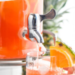 Pinterest graphic for rum punch. Text says "the best rum punch recipe simplejoy.com" Image shows woman's hand holding glass of rum punch with ice and garnished with orange slice and pineapple wedge while drink dispenser pours rum punch into glass.