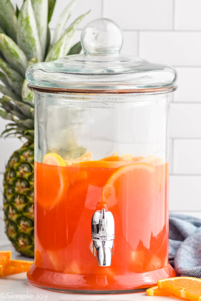 drink dispenser of rum punch with sliced fruit, whole pineapple and orange slices sitting in background