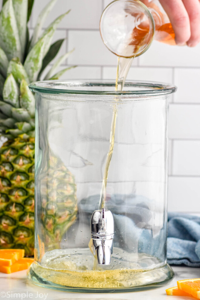 rum pouring into a drink dispenser to make rum punch. Fresh pineapple and orange slices sitting in background